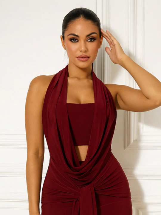 Female model stands before a cream panelled wall. The model wears a slinky red cowl neck halter neck mini dress, with a plunging front and drape design. The model has tanned skin and slick backed brown hair. The model faces forward,  and places one hand on her head and he other by her side. The top of the front can be seen, including the halter plunging cowl front.