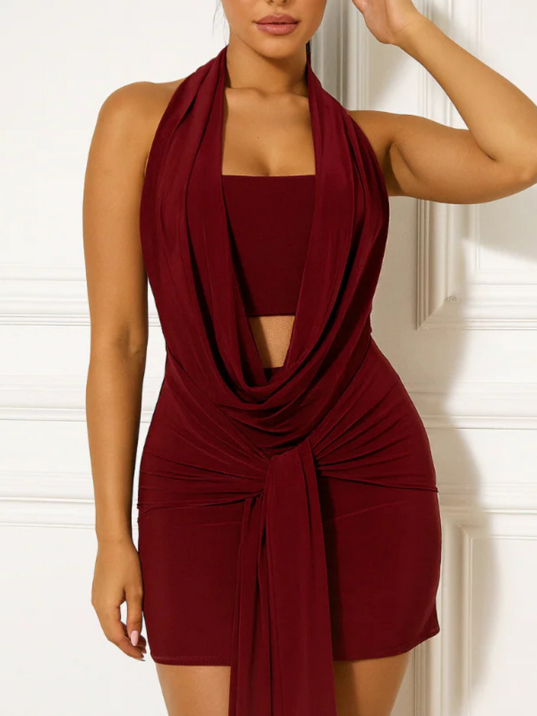 Female model stands before a cream panelled wall. The model wears a slinky red cowl neck halter neck mini dress, with a plunging front and drape design. The model has tanned skin and slick backed brown hair. The model faces forward, the front of the dress can be seen.