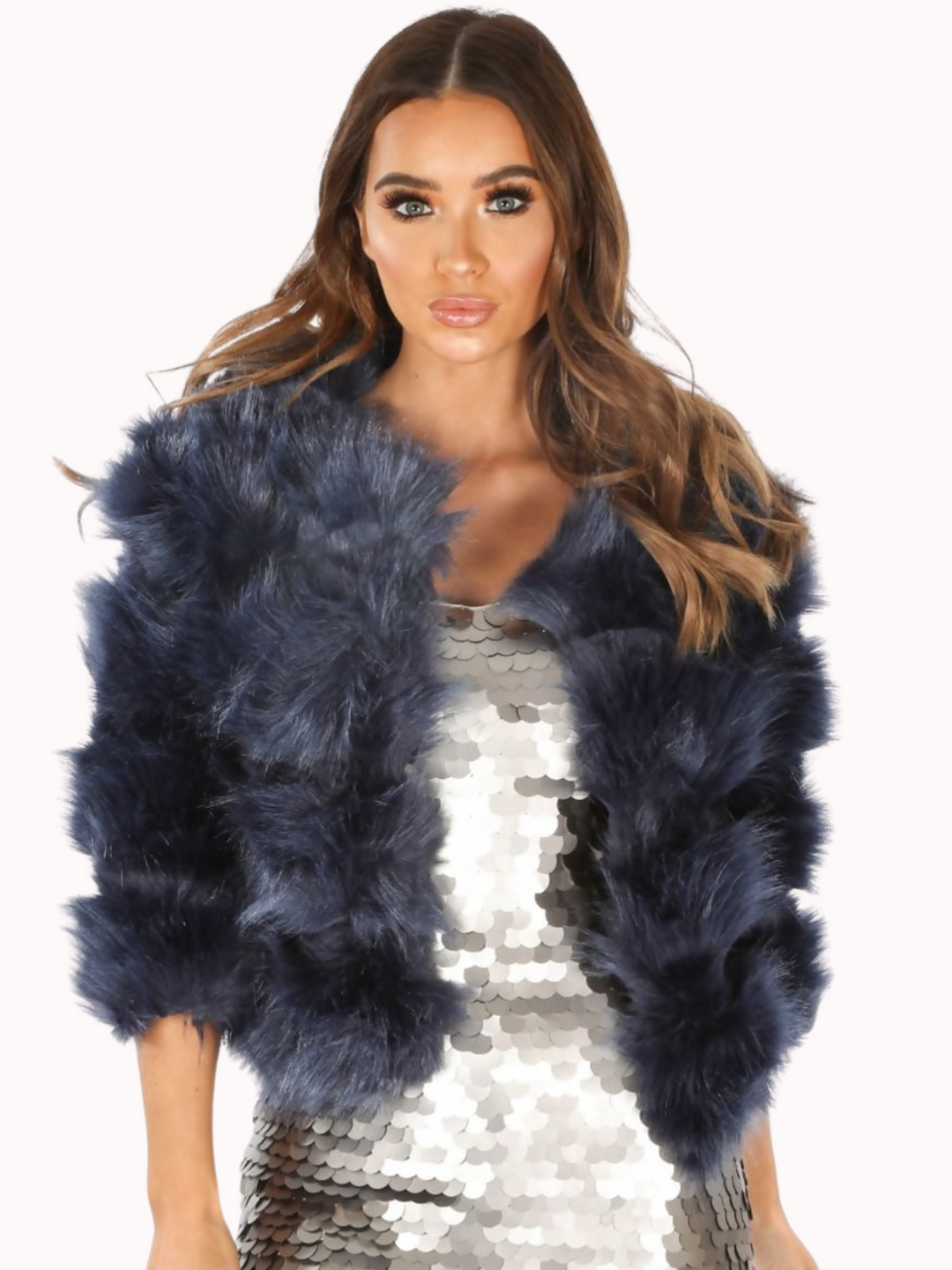 Mid length shot. Model wears a soft faux fur navy jacket with cropped sleeves and front clasp fastening at the top. Model wears  a silver sequin dress underneath the dress and wears the faux fur jacket open. The model faces the camera with both of her hands by her sides.