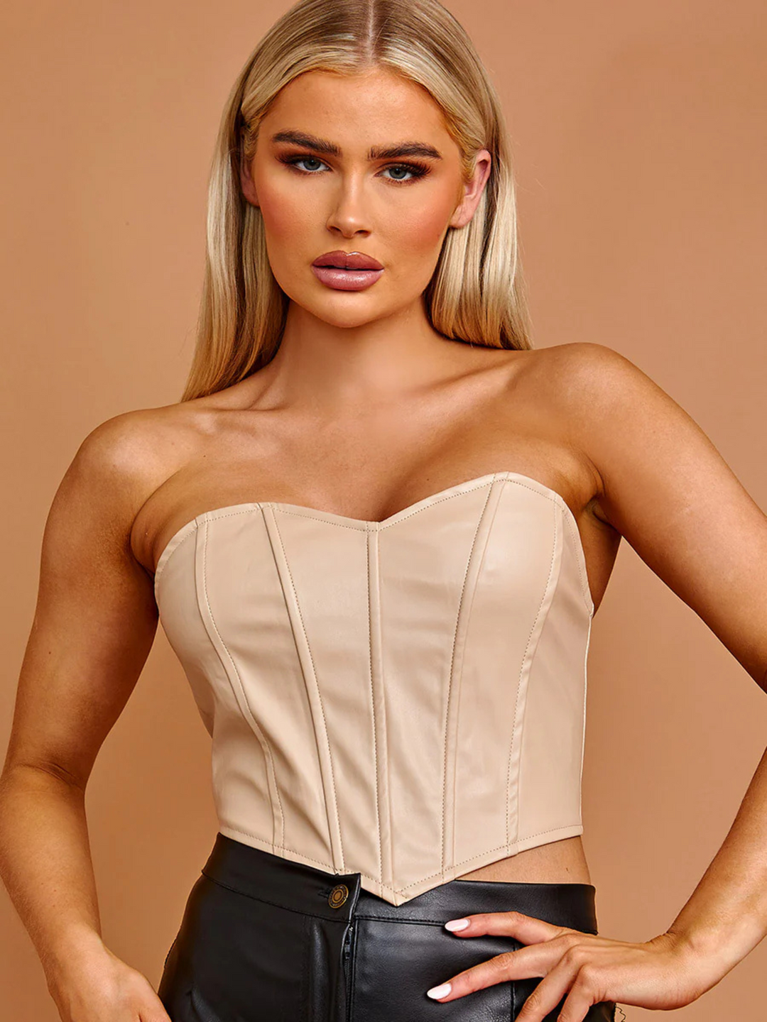 Model wears a stone coloured pu leather bandeau crop top, that has a sweet heart neckline and corset style panellig to the front and zip back fastening to the rear. The model has tanned skin and long blonde hair, she faces the camera placing one hand on her hips and the other by her side.