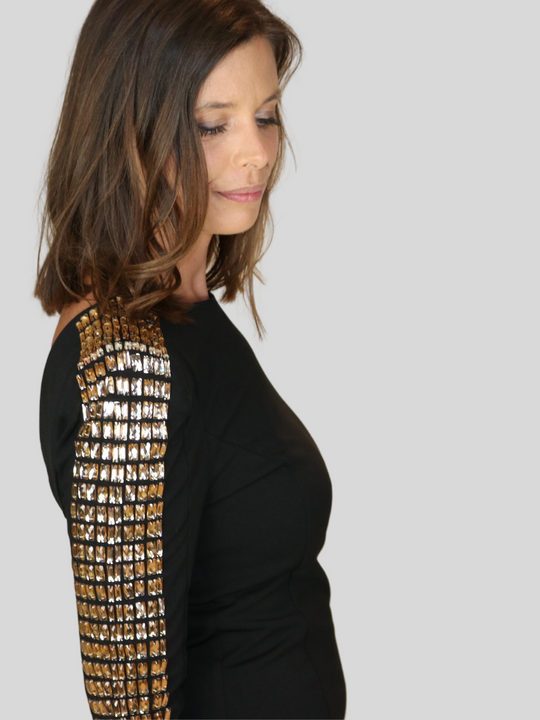 4ever Stunning Deluxe Premium black midi dress with gold rhinestone jeweled arms and chunky gold back zip fastening.  Model stands to the side and looks down on the floor, her jewel sleeved arms can be be seen.