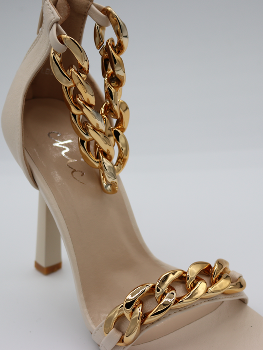A cream shoe with gold detail at the base strap as well as gold chain detailing at the ankle. Shoe placed slightly to the side so that the front and side of the shoe can be seen.