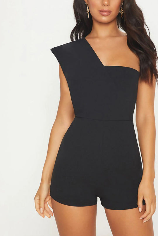 Model wears a black drape one shoulder playsuit. Model faces the camera and has one hand behind her back and one on her leg. Model wears a gold drape earring.