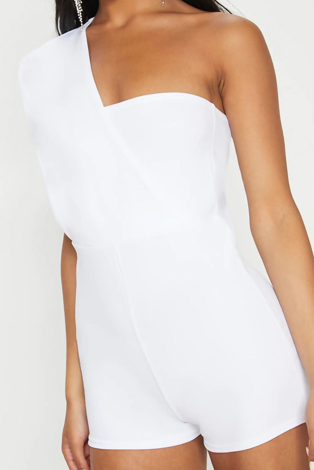 Model wears a white drape one shoulder playsuit. Model stands slightly to the side, where you can see one exposed shoulder and the other shoulder wearing a cross drapped sleeve.
