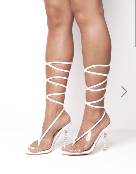 Model wears a diamante heeled shoe. The shoe is in white and has a perspex strap that goes acoss the top of the foot and white straps that criss cross fasten  several times across  the  calf. Model stands with her feet too the side. The side of her shoe showing the diamante  heel is visible.
