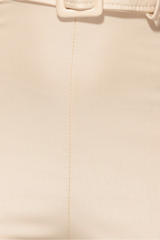 Model wears faux leather beige jumpsuit with high waisted belt . The jumpsuit has a skinny fit  style and a plunging neck line.  Close up of the faux leather material.