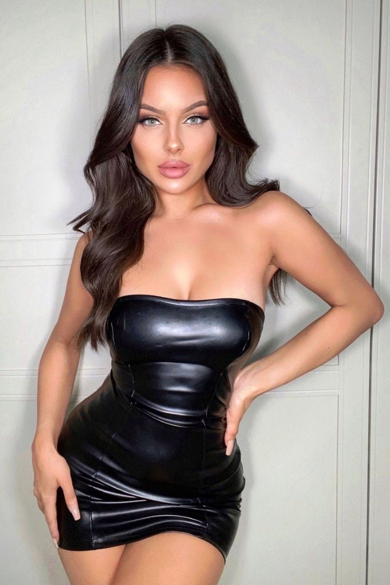 Model wears black faux leather bandeau mini dress. with a bodycon fit. Modek faces the camera and has one leg raised and has one hand on her hip and one touching the the side of her leg. Model stares directly into the camera.