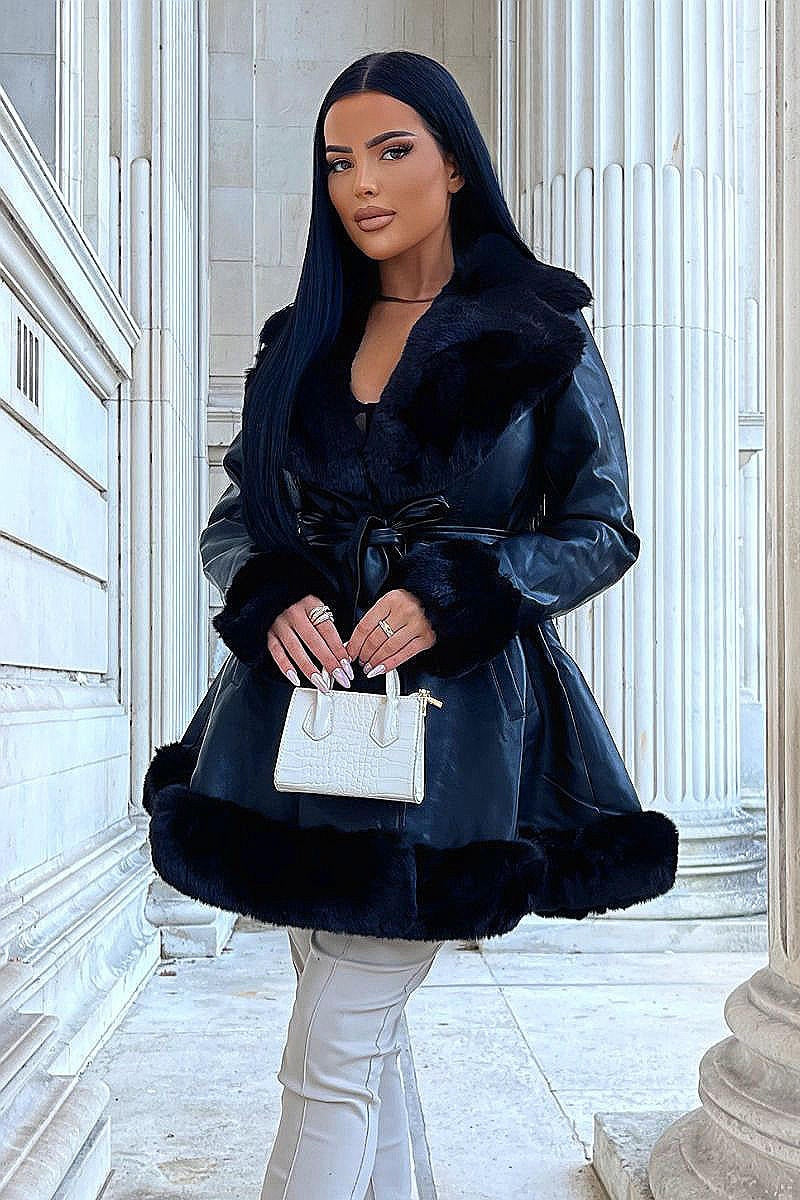 Model wears faux leather swing coat with faux fur tirm at the color, sleeve cuff and the bottom of the coat. The coat has a central button fastening and a faux leather belt. Model is outside and faces the camera with her legs crossed. Model holds a small top handle hand bag which she holds with both hands.