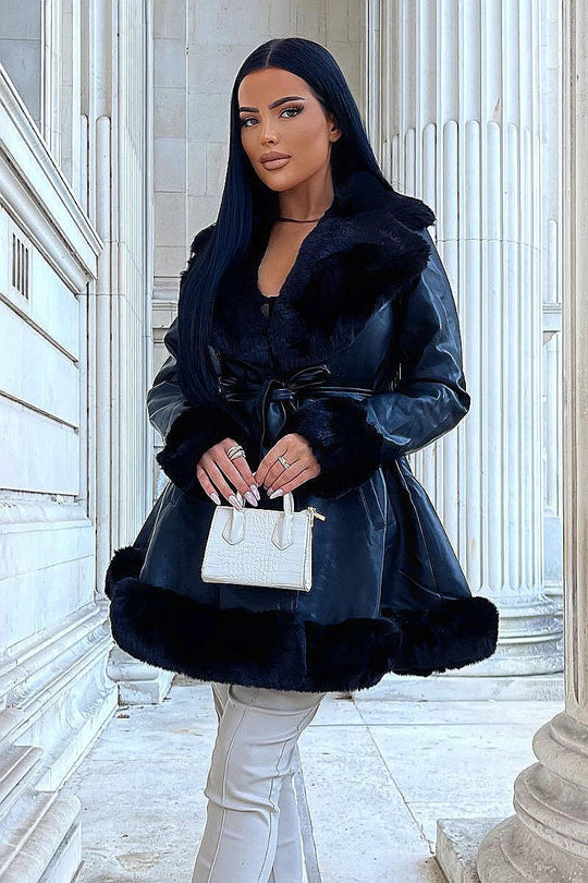 Model wears faux leather swing coat with faux fur tirm at the color, sleeve cuff and the bottom of the coat. The coat has a central button fastening and a faux leather belt. Model is outside and faces the camera with her legs crossed. Model holds a small top handle hand bag which she holds with both hands.