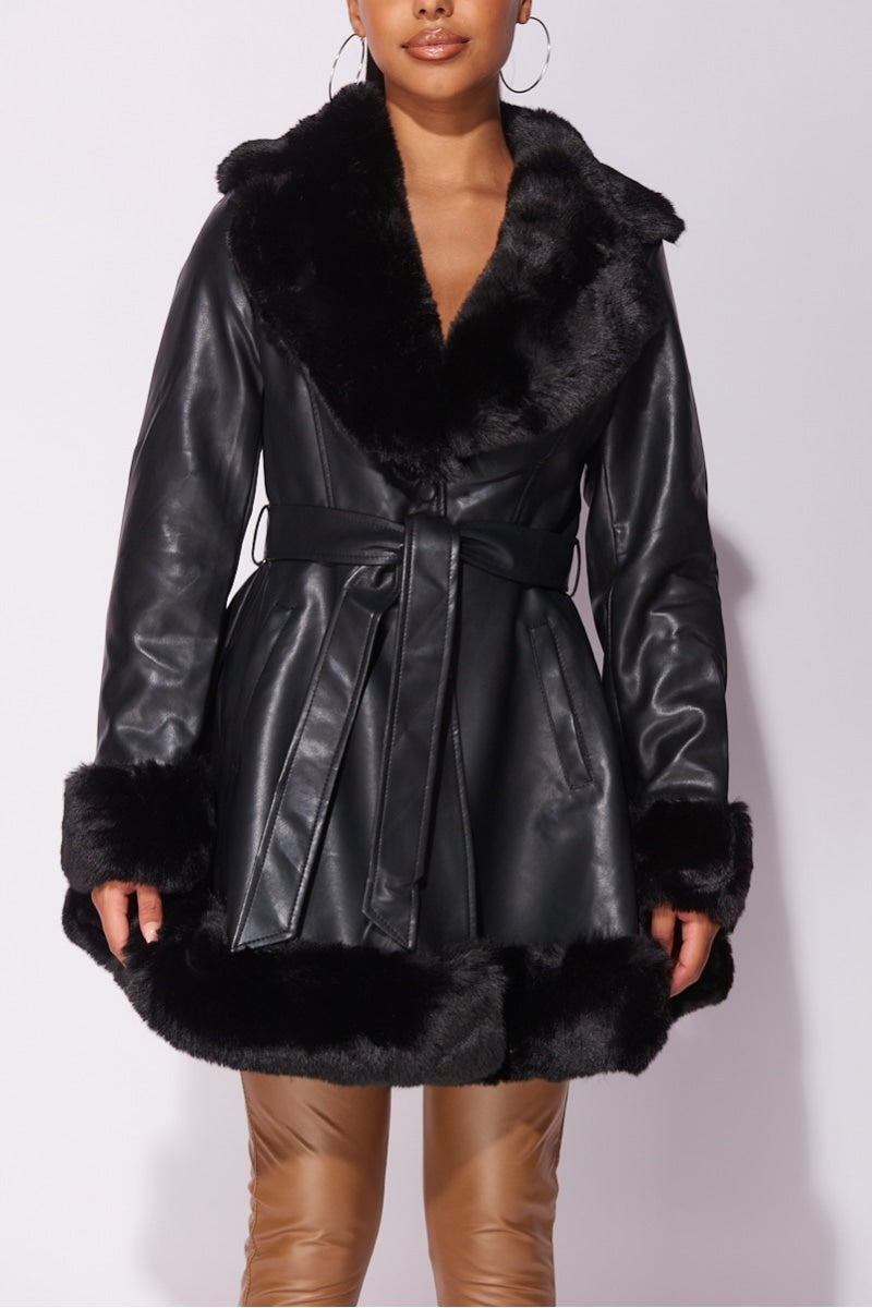 Model wears faux leather swing coat with faux fur tirm at the color, sleeve cuff and the bottom of the coat. The coat has a central button fastening and a faux leather belt. Model wears brown faux leather jeggings. Model has her hands to the side.