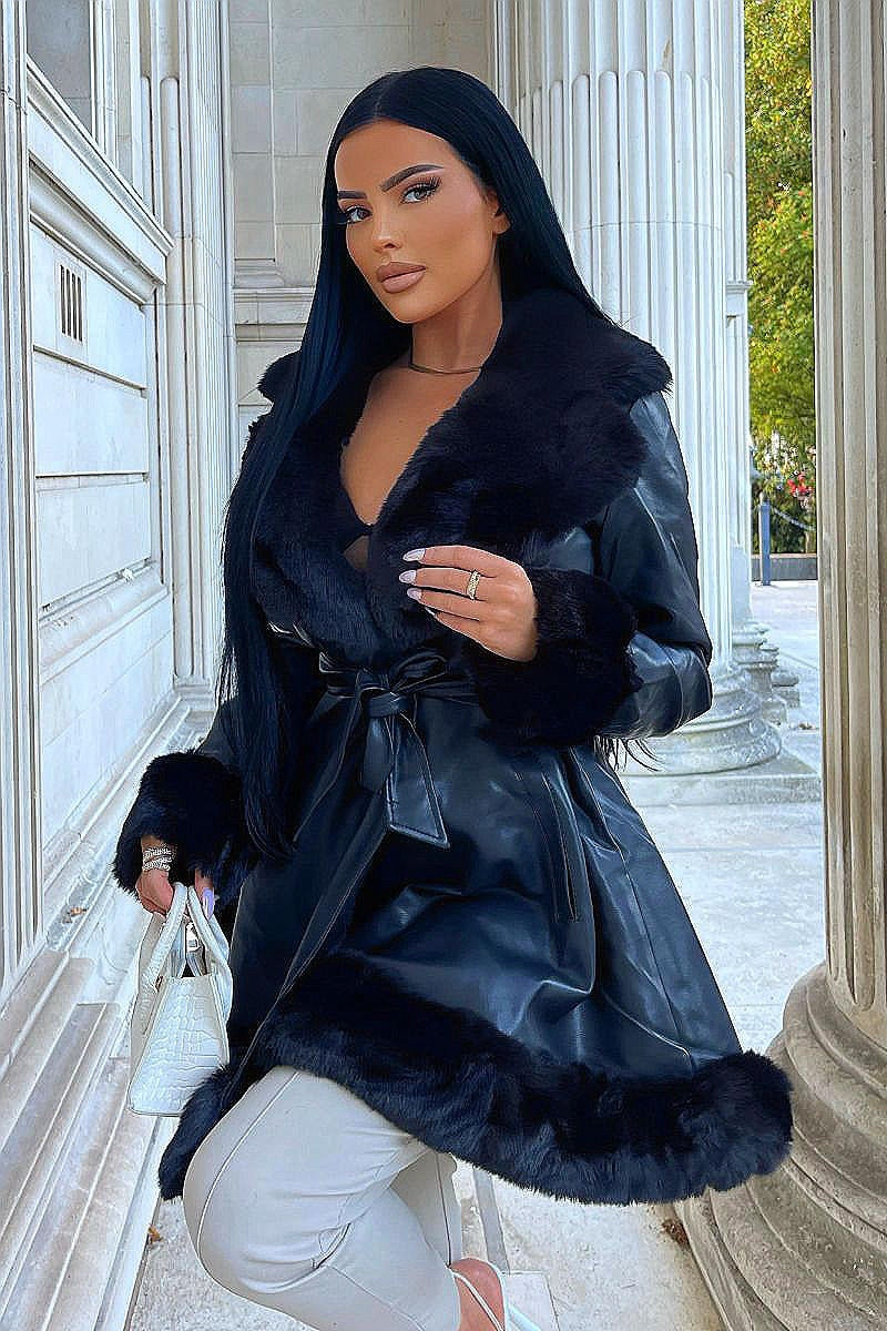 Model wears faux leather swing coat with faux fur tirm at the color, sleeve cuff and the bottom of the coat. The coat has a central button fastening and a faux leather belt. Model is outside. Model raises one leg  and holds a small bag with the other.