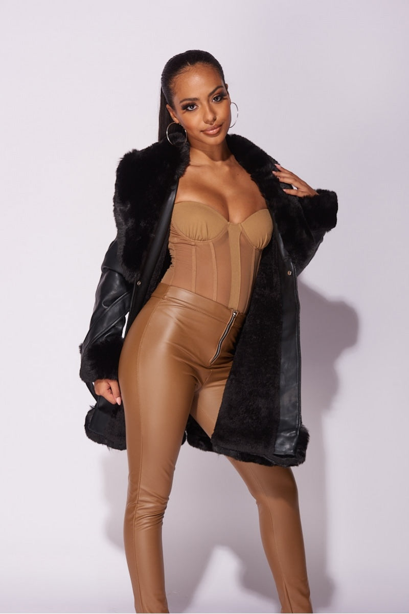 Model wears faux leather swing coat with faux fur tirm at the color, sleeve cuff and the bottom of the coat. The coat has a central button fastening and a faux leather belt. Model wears a beige bodysuit and faux leather jeggings. Model has the coat open with one side of the coat behind he back and one at teh front.
