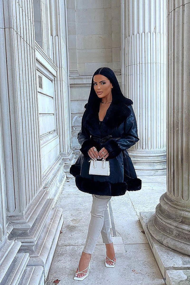 Model wears faux leather swing coat with faux fur tirm at the color, sleeve cuff and the bottom of the coat. The coat has a central button fastening and a faux leather belt. Model is outside. Model  stands with one leg in front of the other. Model holds a small bag  and is wearing faux leather jeggings and strappy heels.
