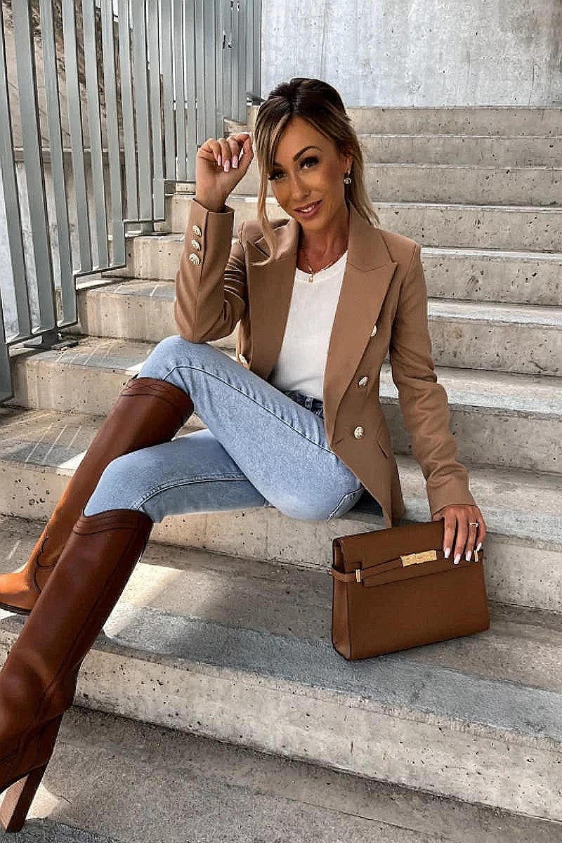 Model wears a camel coloured military button inspred blazer with padded shoulders and button fastening. Model sits on a step and looks up. Model holds a brown clutch bag and matching shoes. Model  is wearing light blue denim jeans.