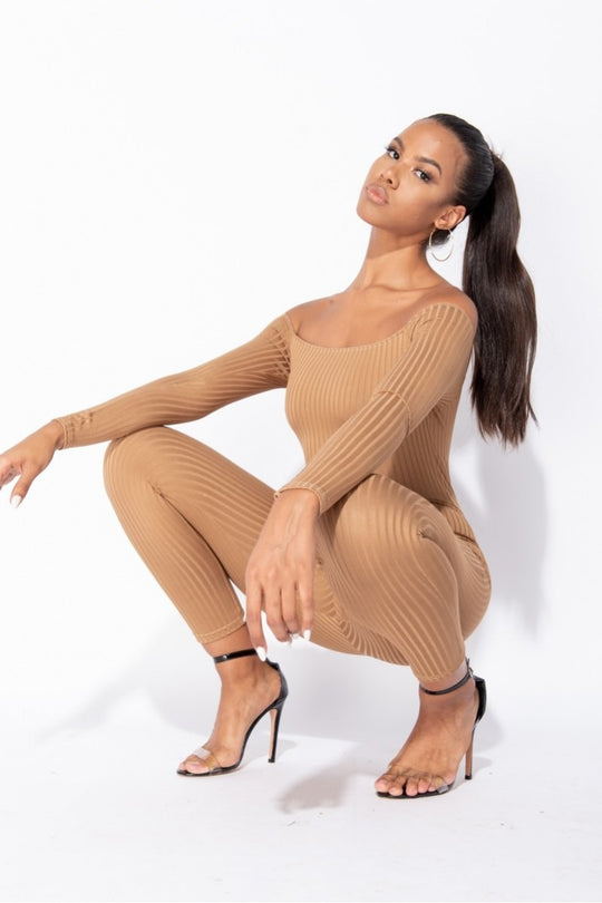Model wears camel coloured ribbed jumpsuit with bardot neckline. Model crouches down and is wearing black strappy heels. Model tilts her head and stares directly into the camera.