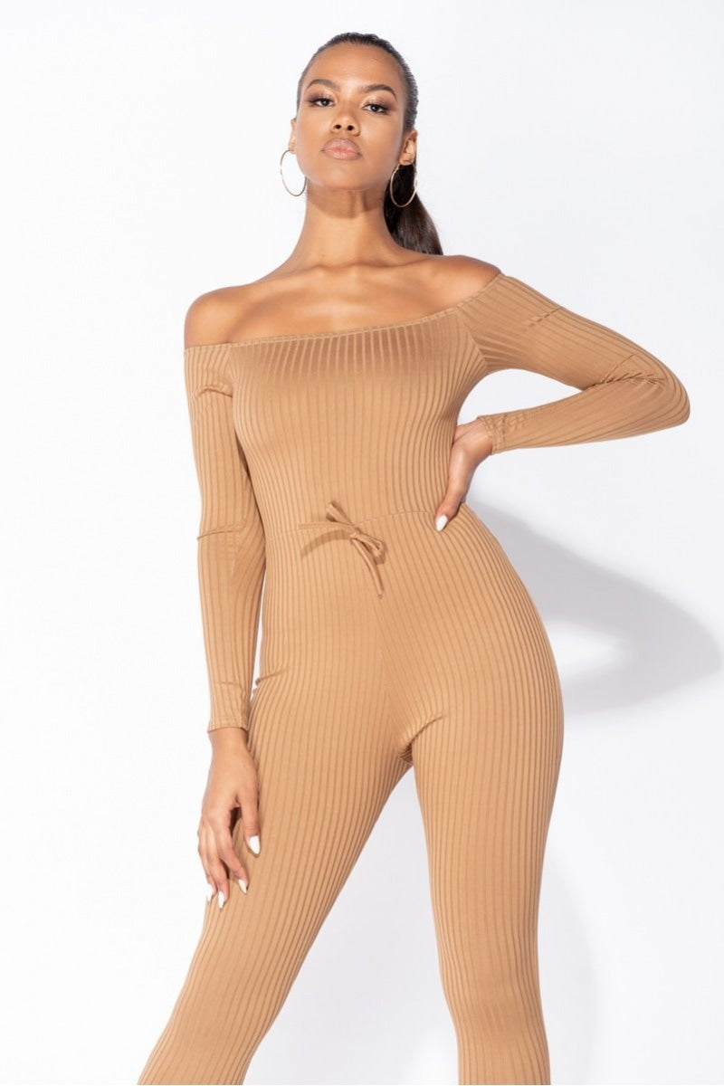 Model wears camel coloured ribbed jumpsuit with bardot neckline.  Model looks into the camera with one hand on her hip and the other touching her leg.