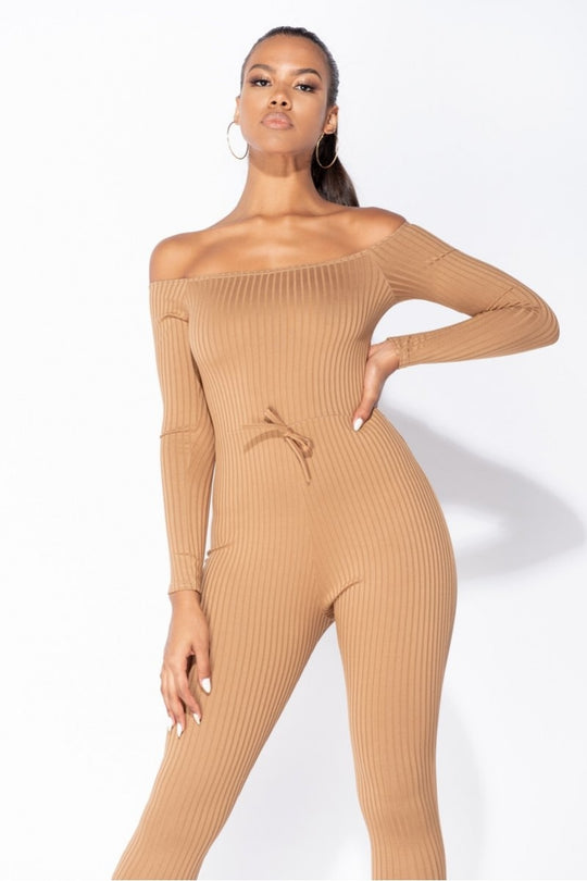 Model wears camel coloured ribbed jumpsuit with bardot neckline.  Model looks into the camera with one hand on her hip and the other touching her leg.