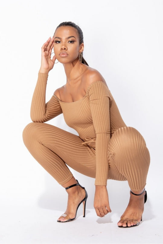 Model wears camel coloured ribbed jumpsuit with bardot neckline.  Model crouches down, with one  arm at the side of her leg and the other hand touching her face. Model stares directly into the camera