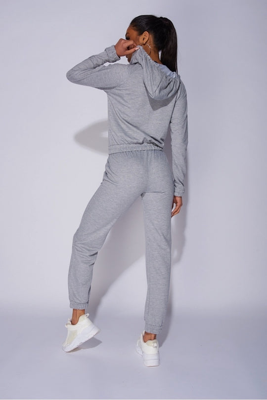 Model wears a light weight tracksuit with front zip fastening and draws string hood. Model places her back too the camera. The back of the tracksuit is visible, including the hood which the model holds with one hand.