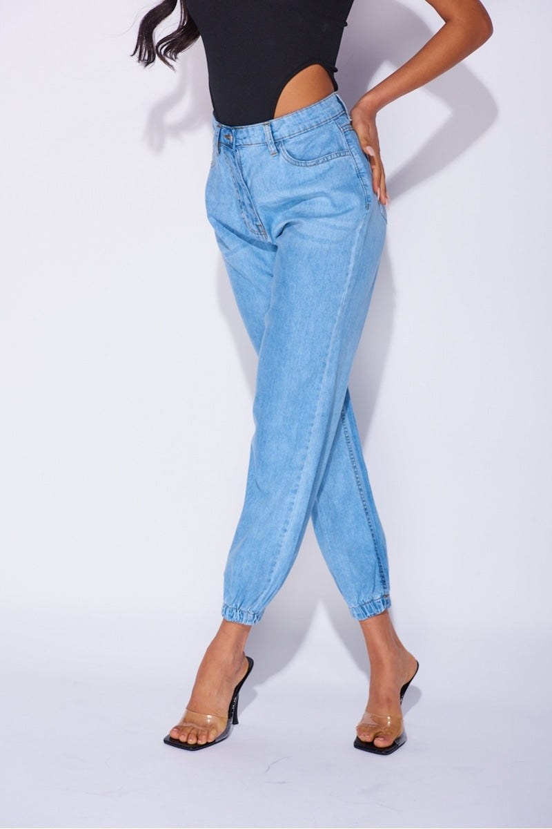 Model wears blue ciffed denim joggers with front zip fastening. Model wears a black high rise body suit.  Model stands with her legs crossed and  one hand on her hip.