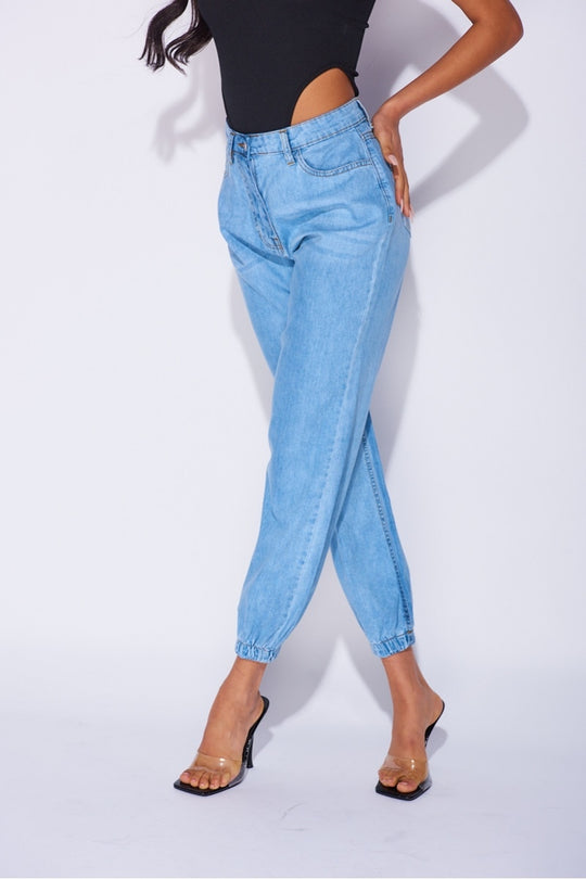 Model wears blue ciffed denim joggers with front zip fastening. Model wears a black high rise body suit.  Model stands with her legs crossed and  one hand on her hip.
