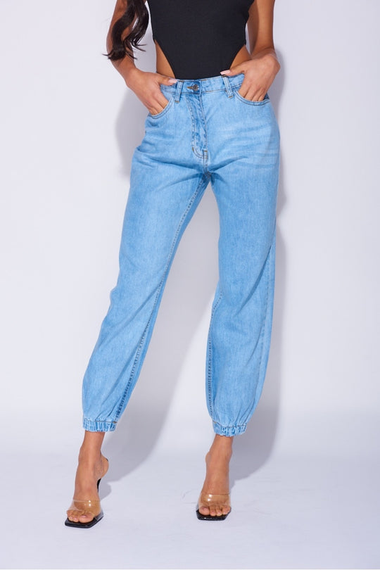 Model wears blue ciffed denim joggers with front zip fastening. Model wears a black high rise body suit. Model places her hands in the front pockets.