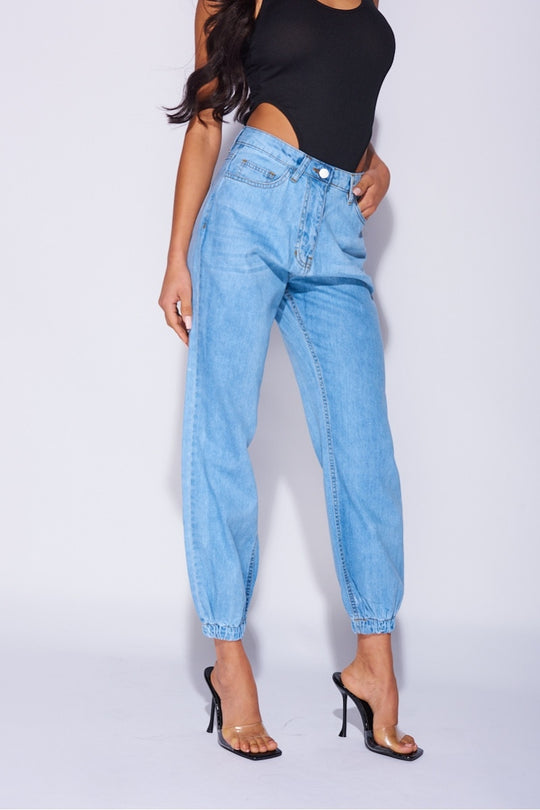 Model wears blue ciffed denim joggers with front zip fastening. Model wears a black high rise body suit. Model stands to the side. the front of the jeans is visible.