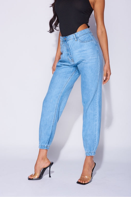Model wears blue ciffed denim joggers with front zip fastening. Model wears a black high rise body suit.  Model stands to the side with one leg in front of the other and her hands by her sides.