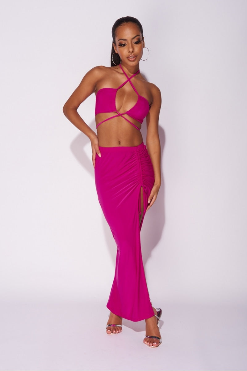 Model wears a magenta co-ord set. The top has a plunging criss cross crop top and the skirt has a thigh split and length adjusting string.   Model places one hand by her side and the other on her hip.