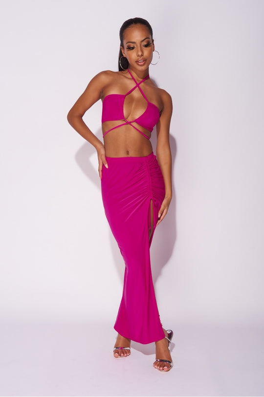 Model wears a magenta co-ord set. The top has a plunging criss cross crop top and the skirt has a thigh split and length adjusting string.   Model places one hand by her side and the other on her hip.