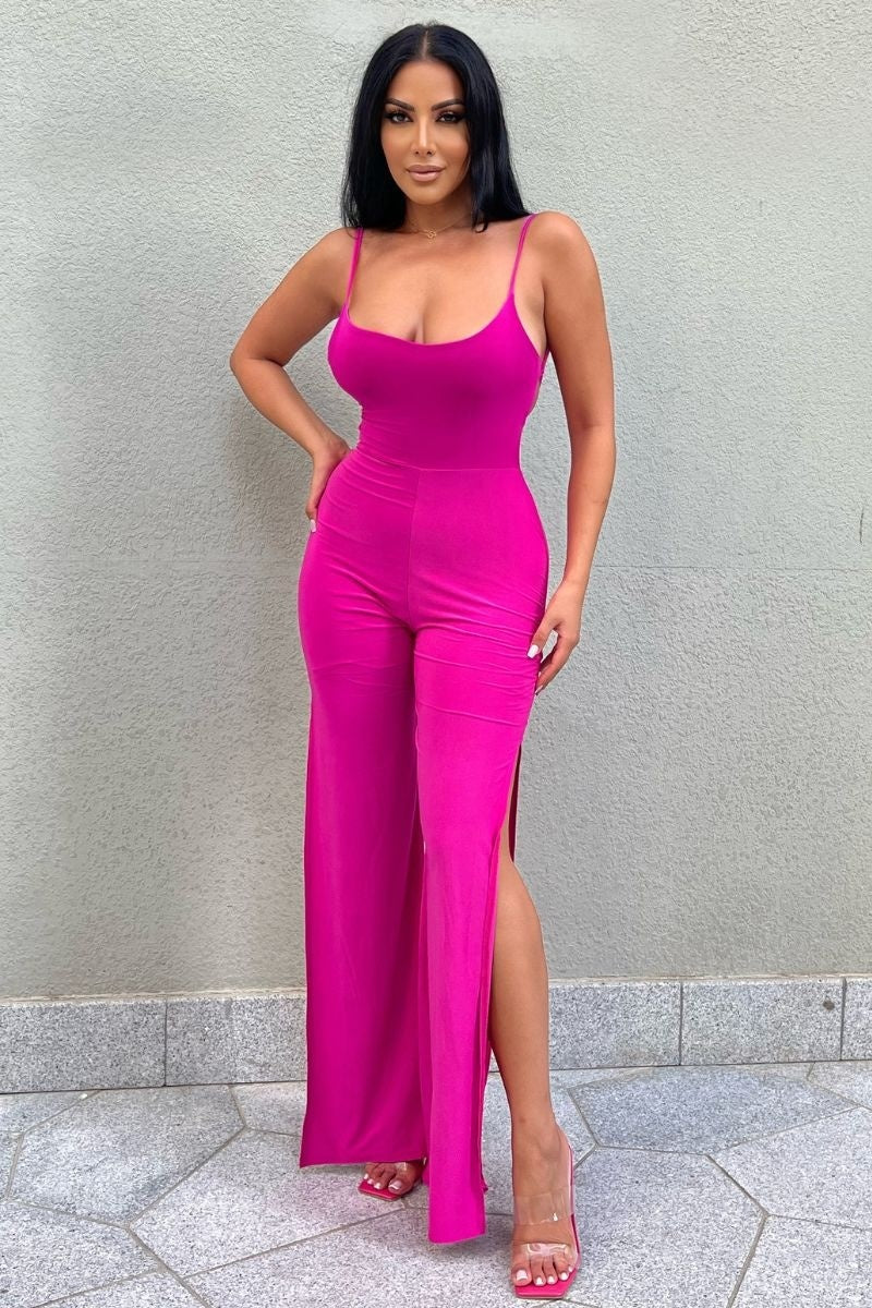 Model wears slinky magenta jumpsuit with a  scoop neck and spagehetti strap.  The jumpsuit  has thigh a thigh high split on each leg.  Model stands with one hand on her hip and the other by her side.