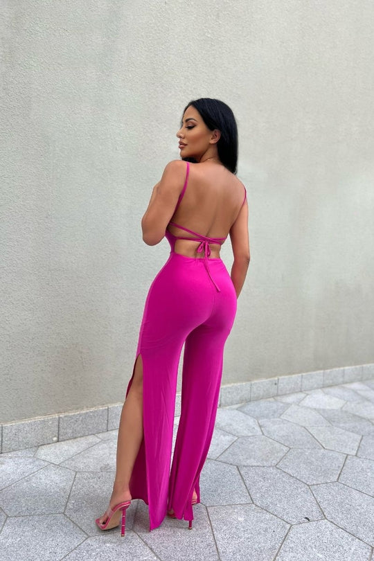 Model wears slinky magenta jumpsuit with a  scoop neck and spagehetti strap.  The jumpsuit  has thigh a thigh high split on each leg.  The jumpsuit has a low back and strap fastening. Model  stands with her back showing, demonstrating the low back jumpsuit.