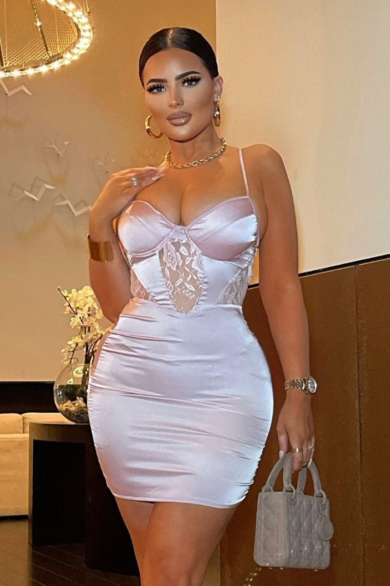 Model wears pink satin dress with front lace panel detailing with cup detailing. Model holds a bag and places one  hand on her collar bone.