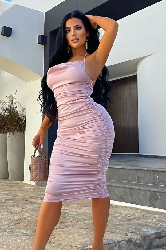 Model wears pink satin midi dress, with a cowl neck and thinned straped halter neck detailing. The dress has ruched detailing at the front and rear of the dress. Model holds a bag in one hand and her hand above her head with the other.