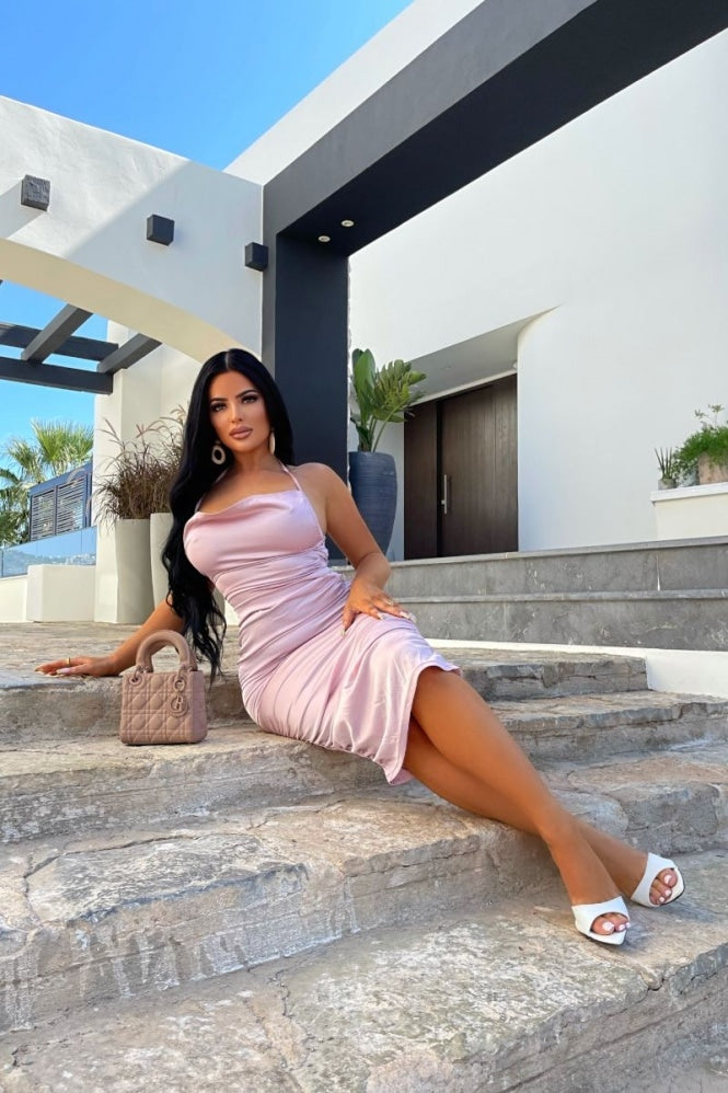 Model wears pink satin midi dress, with a cowl neck and thinned straped halter neck detailing. The dress has ruched detailing at the front and rear of the dress.  Model sits on the step outside with her legs crossed. Model plaes one hand on her leg and the other on the step.