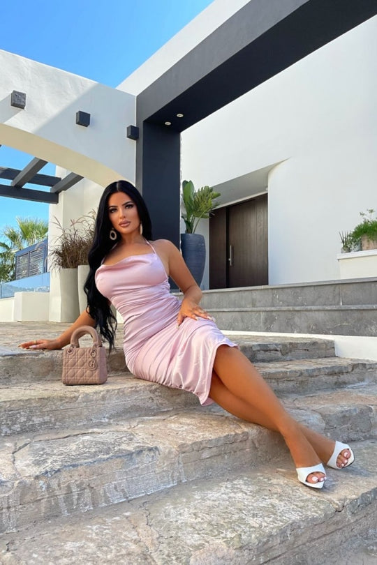 Model wears pink satin midi dress, with a cowl neck and thinned straped halter neck detailing. The dress has ruched detailing at the front and rear of the dress.  Model sits on the step outside with her legs crossed. Model plaes one hand on her leg and the other on the step.