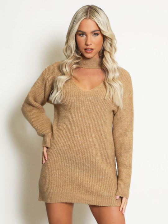 Female model stands in front of a plain background. The model wears a beige knitted long sleeved mini jumper dress. The jumper dress has a high neck and cut out front. The model has tanned skin and wavy blonde hair, she faces the camera and places one hand on her hips and the other by her side.