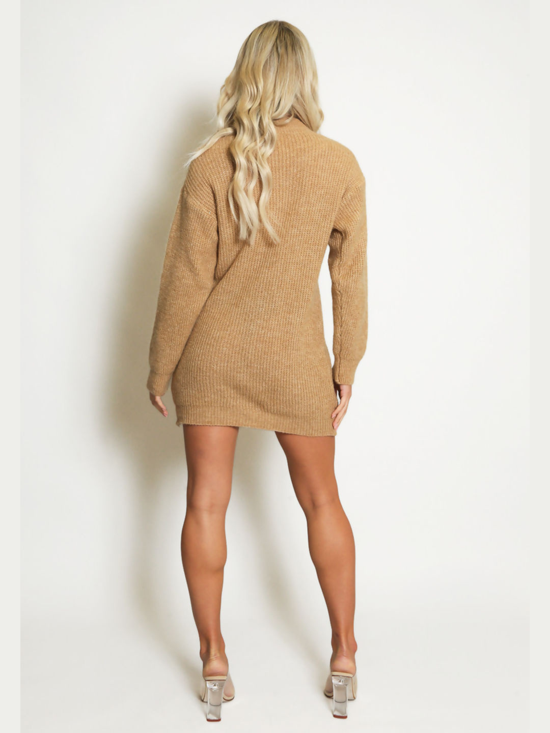 Full length shot. Female model stands in front of a plain background. The model wears a beige knitted long sleeved mini jumper dress. The jumper dress has a high neck and cut out front. The model has tanned skin and wavy blonde hair, she stands  with her back to the camera, the back of the jumper dress can be seen, she wears perspex high heeled shoes 