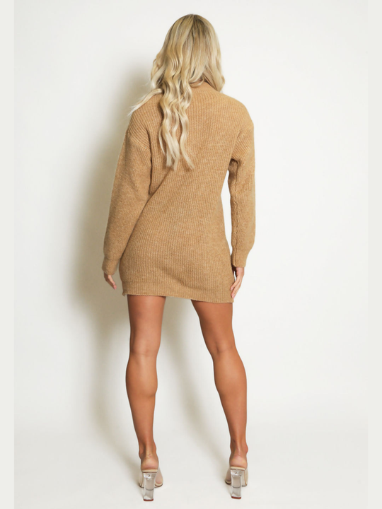 Full length shot. Female model stands in front of a plain background. The model wears a beige knitted long sleeved mini jumper dress. The jumper dress has a high neck and cut out front. The model has tanned skin and wavy blonde hair, she stands  with her back to the camera, the back of the jumper dress can be seen, she wears perspex high heeled shoes 