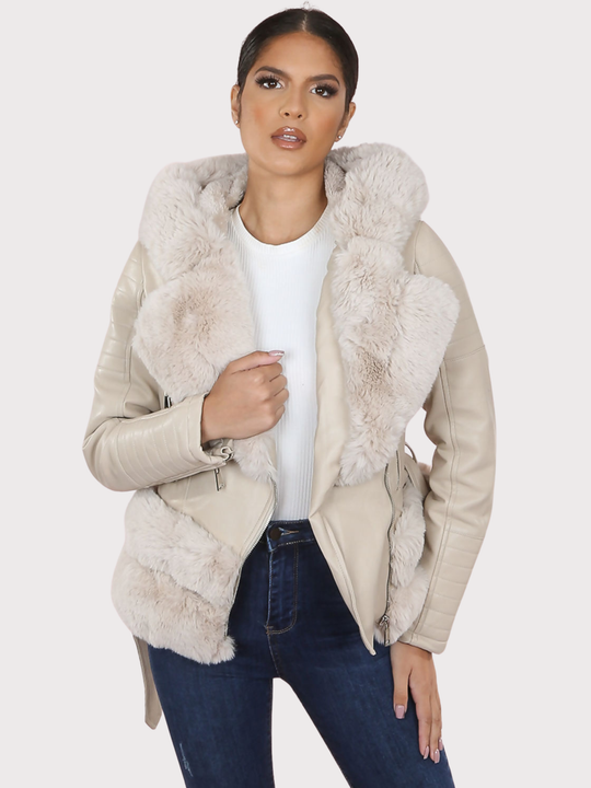 Female model stands in front of a plain background. The model wears a beige pu leather biker jacket with belted waist, zip front fastening and beige faux fur lapel and beige faux fur trim. The model faces the camera, the front of the jacket can be seen, including he faux fur trim at the base of the jacket.  The model has tanned skin and dark brown hair, she faces the camera with the jacket open. The model places one hand on the faux fur lapel and the other on her thigh. 