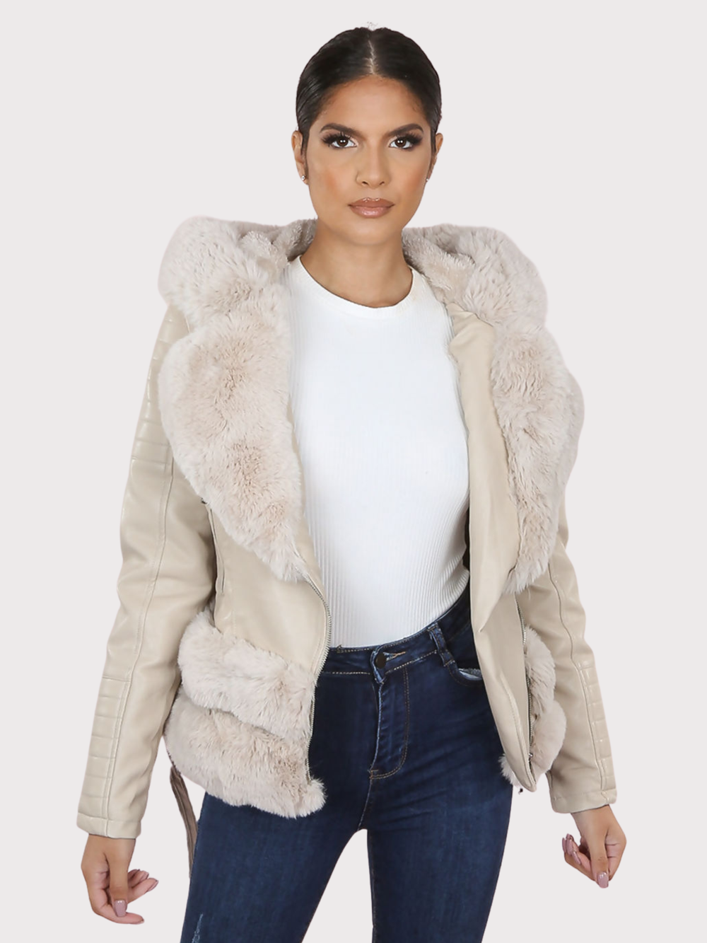 Female model stands in front of a plain background. The model wears a beige pu leather biker jacket with belted waist, zip front fastening and beige faux fur lapel and beige faux fur trim. The model faces the camera, the front of the jacket can be seen, including he faux fur trim at the base of the jacket.  The model has tanned skin and dark brown hair, she stands faces the camera with her jacket open and her hands by her sides. 