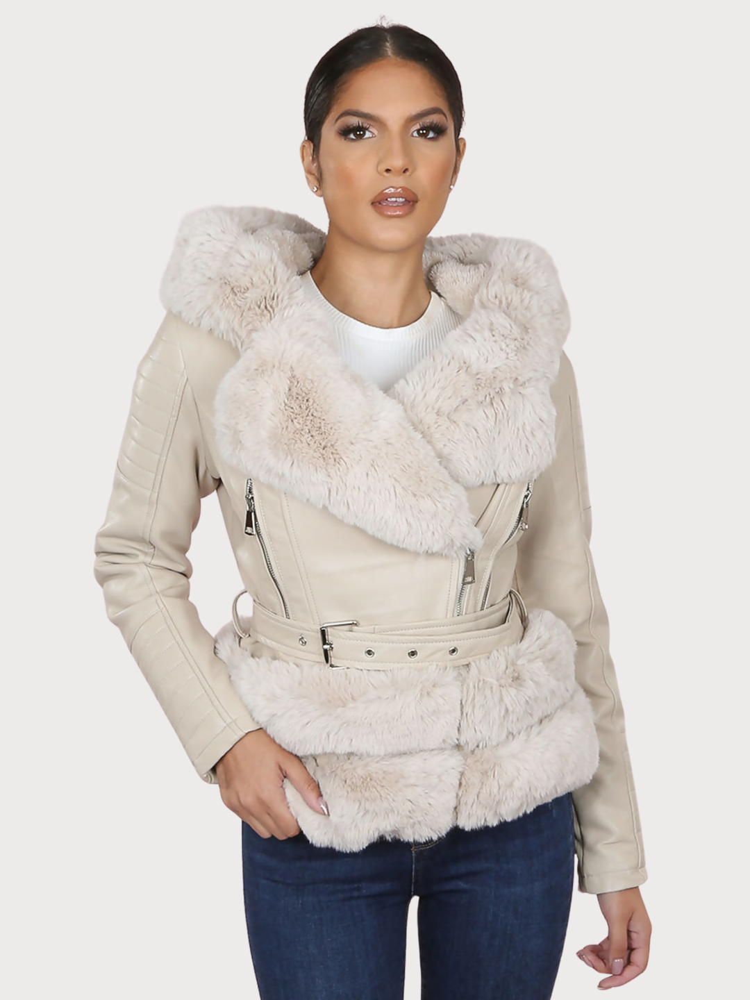 Female model stands in front of a plain background. The model wears a beige pu leather biker jacket with belted waist, zip front fastening and beige faux fur lapel and beige faux fur trim. The model faces the camera, the front of the jacket can be seen, including he faux fur trim at the base of the jacket.  The model has tanned skin and dark brown hair. The model faces the camera, she places one hand on the faux fur trim at the base of the jacket &  one hand by her side, she looks directly into the camera. 