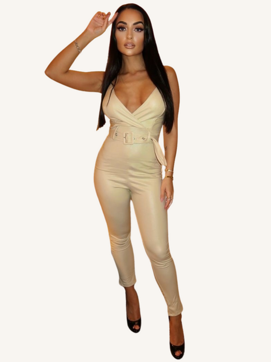 Model wears a beige faux leather jumpsuit with belted waist and plunging neckline. Model faces forward and puts one hand to her head.