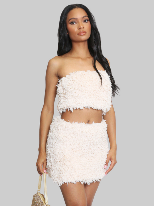 Woman wearing a beige textured two-piece outfit on a white background. The co-ordinated set has a shaggy texture with silver flex detailing, the shaggy bandeau crop top has a matching mini skirt. The model has light brown skin and long black hair, she faces the camera and places both hands by her sides.  The model wears silver holds a dressy  gold hand bag. 