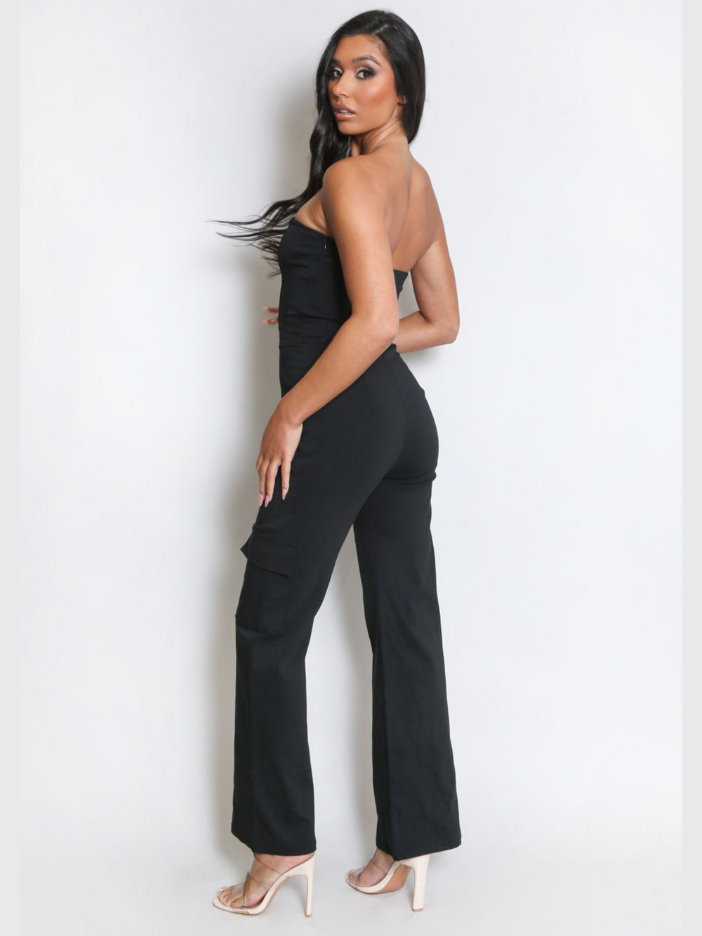 Full length. Female model wears a black jumpsuit that has cargo pockets, is strapless and has a bandeau neckline. The model has tanned skin and long black hair, she stands to the side, placing one hand on her thigh, she turns to look into the camera; the thigh cargo pocket can be seen. The model wears high heels. 