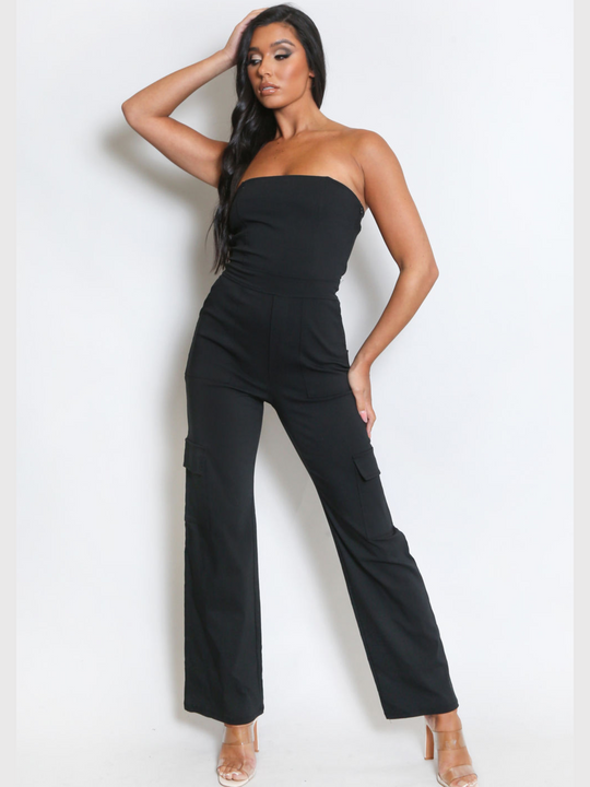 Female model wears a black jumpsuit that has cargo pockets, is strapless and has a bandeau neckline. The model has tanned skin and long black hair, she faces the camera and looks off to the side. The model places one hand on her head and the other at her thigh, she wears perspex high heels. 