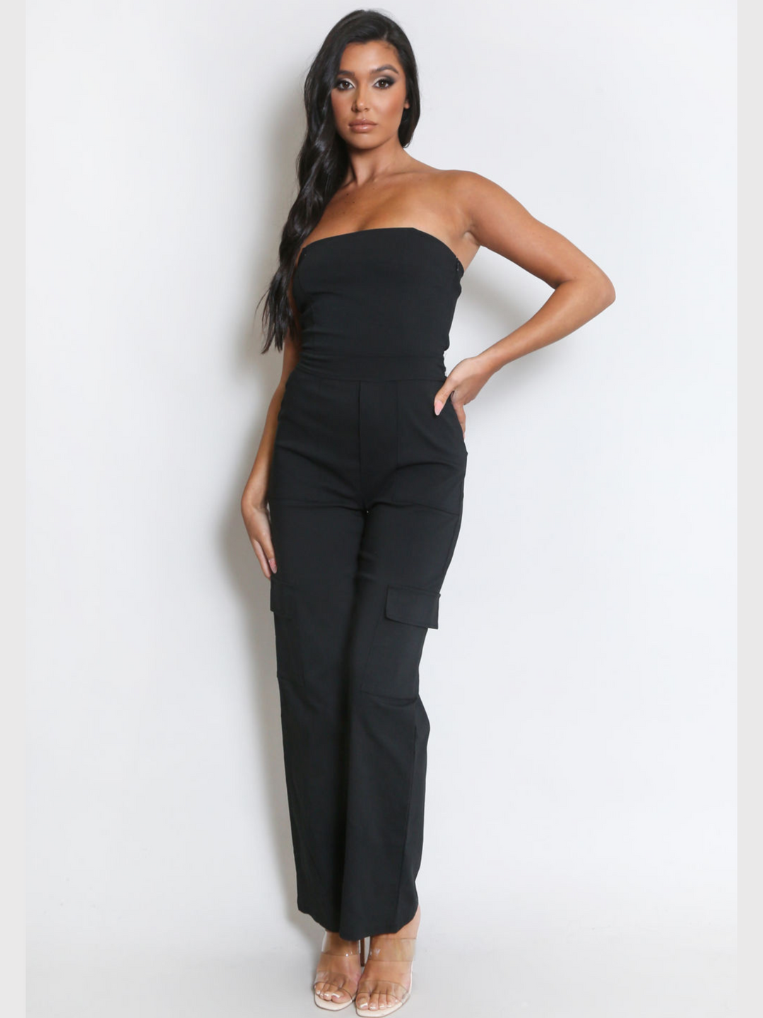 Full length shot. Female model wears a black jumpsuit that has cargo pockets, is strapless and has a bandeau neckline. The model has tanned skin and long black hair, she faces the camera, placing one hand on her hip and the other by her side. 