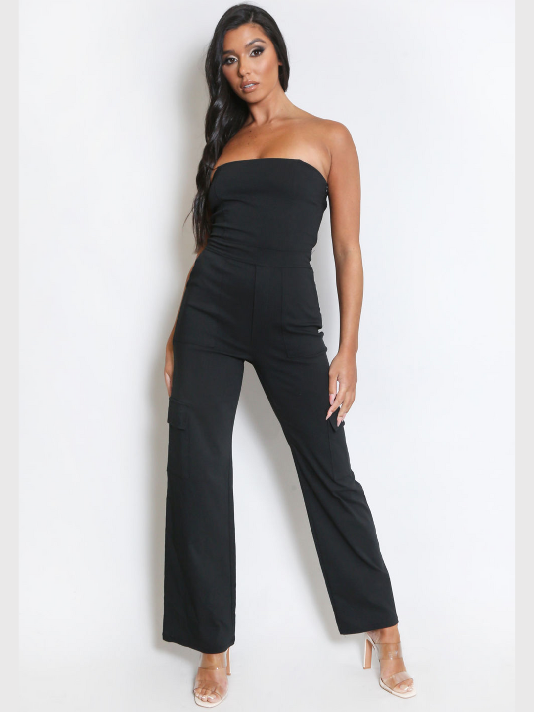 Full length shot. Female model wears a black jumpsuit that has cargo pockets, is strapless and has a bandeau neckline. The model has tanned skin and long black hair, she faces the camera placing both hands by her sides, she wears clear perspex heels. 