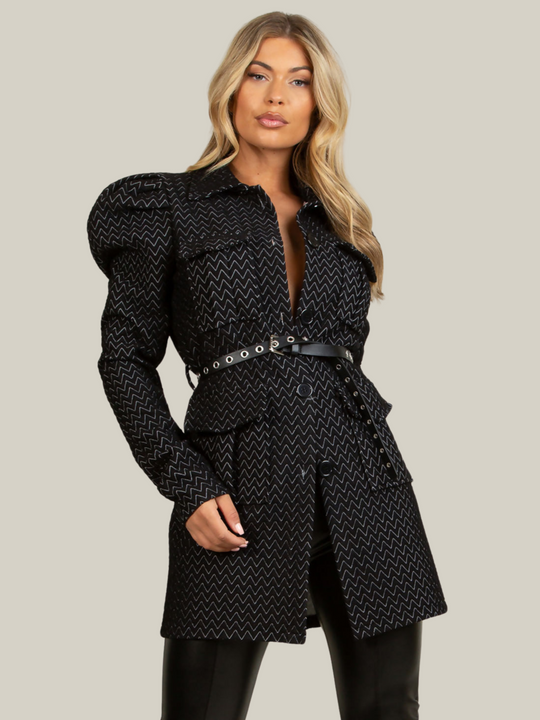 Model wears a black longline blazer with soft quilted patterns with a soft textured finish. The shoulders have a puffed finish with long sleeves and front button fastening. The model wears a belt that  is fed between the belt loop. Model faces the camera with one hand by her side and one hand behind her back.  