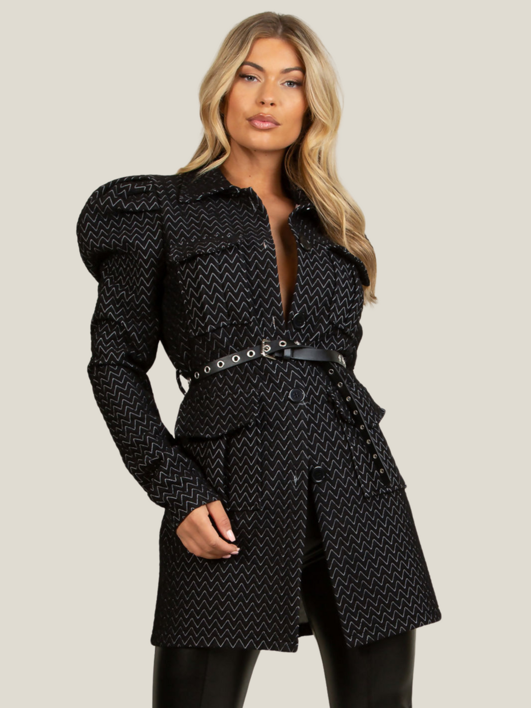Model wears a black longline blazer with soft quilted patterns with a soft textured finish. The shoulders have a puffed finish with long sleeves and front button fastening. The model wears a belt that  is fed between the belt loop.  Model stands with one hand behind her back. 